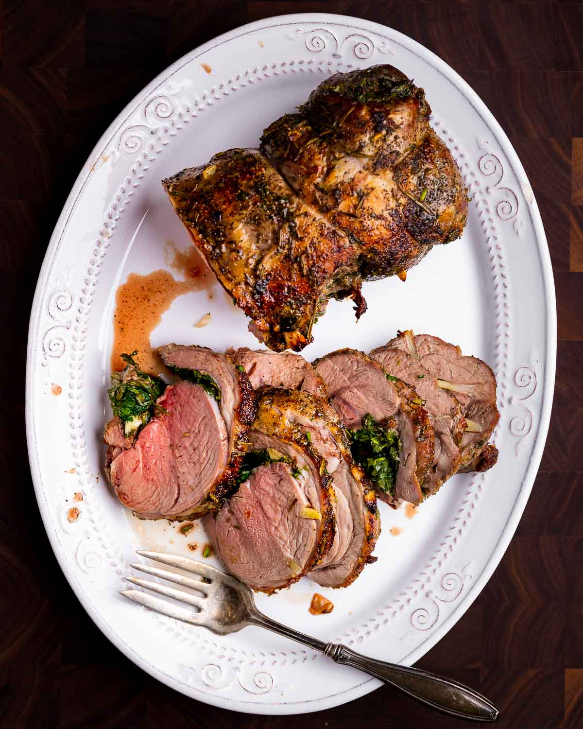 Sliced leg of lamb in white platter on brown cutting board.