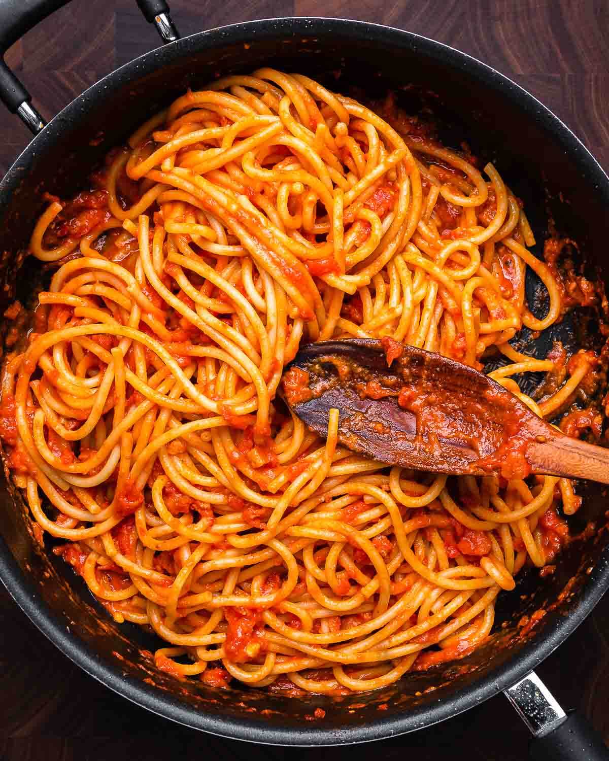 Ovehead shot of large pan with bucatini Amatriciana and wooden spoon.