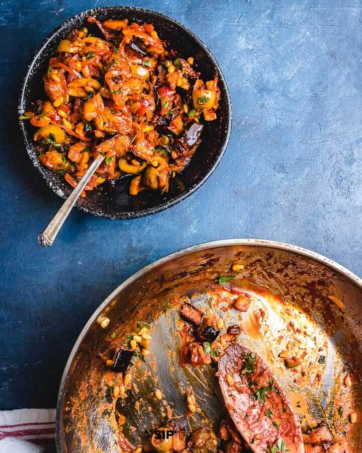 Large pan and small black bowl of caponata on blue table.