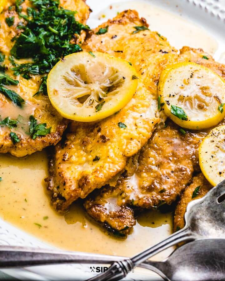 Chicken Francese in white platter with lemons and parsley on top.