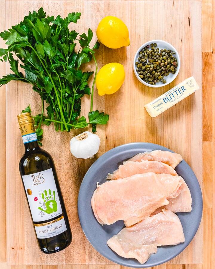 Ingredients shown on cutting board: parsley, lemons, capers, butter, wine, garlic, and chicken cutlets.