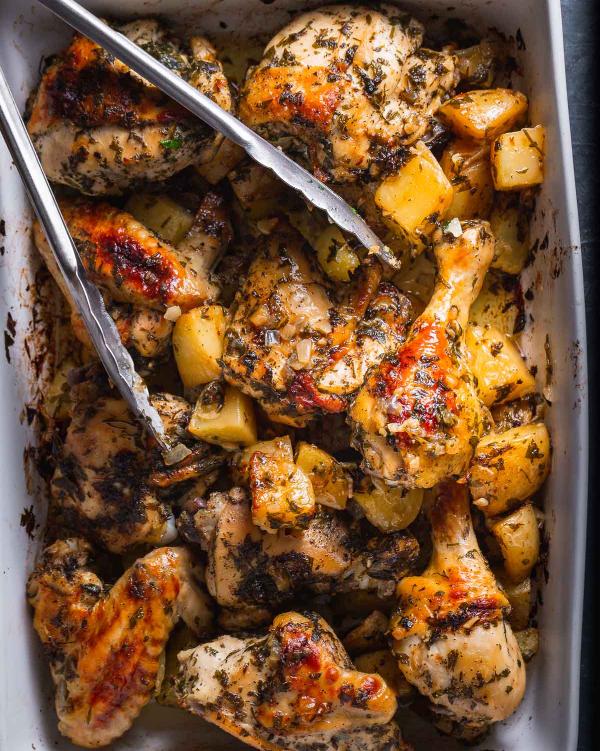 Overhead shot of Italian baked chicken and potatoes in white baking dish with tongs.