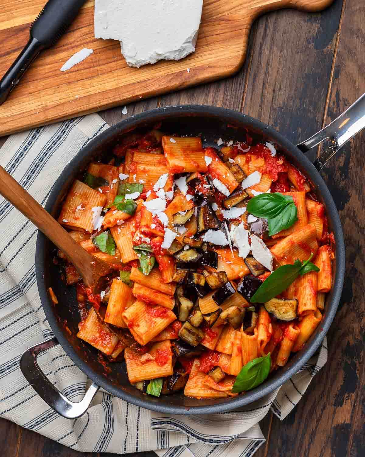 Overhead shot of large pan with pasta alla Norma and block of ricotta salata on the side.