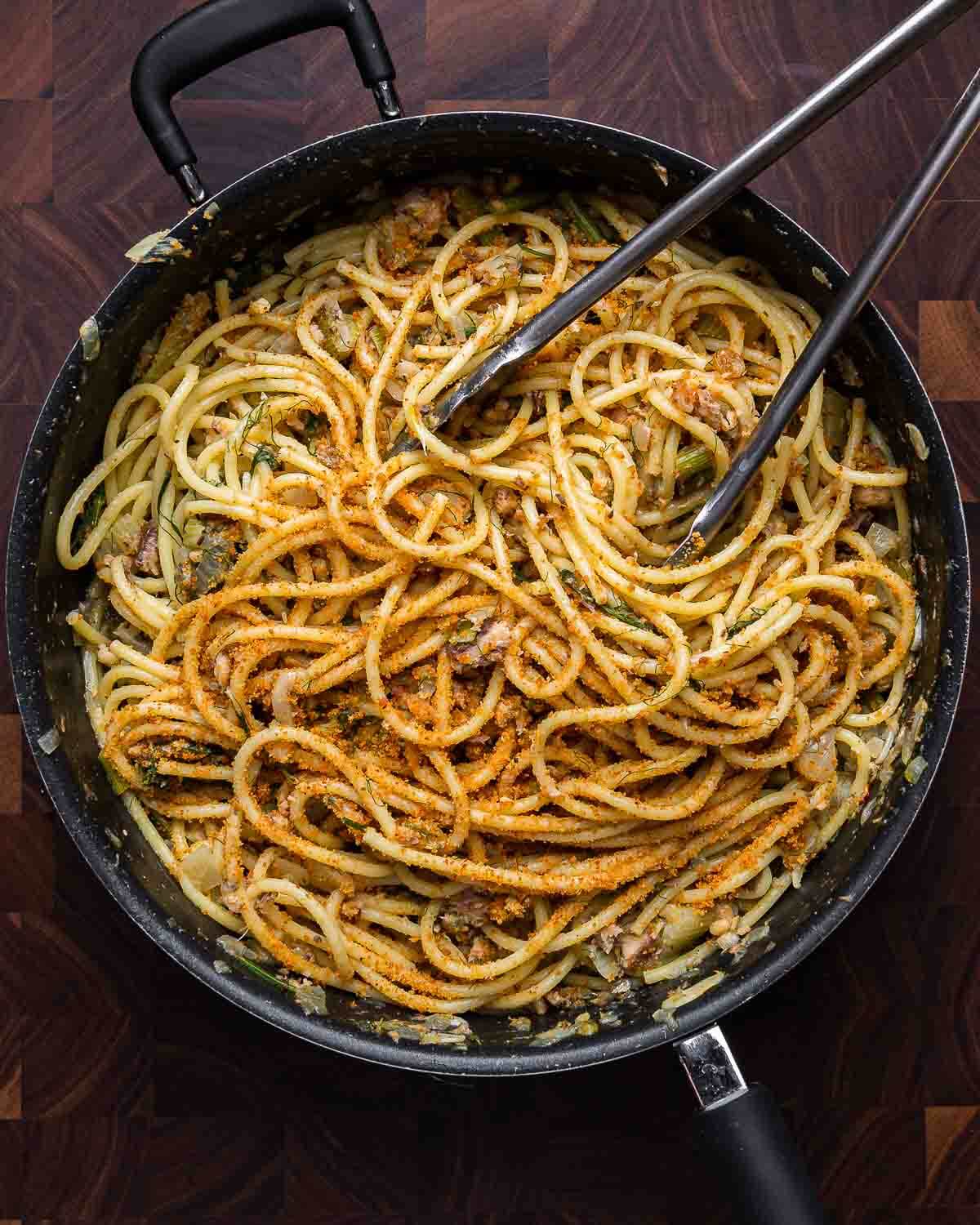 Overhead shot of pasta con le sarde in black pan on walnut cutting board.