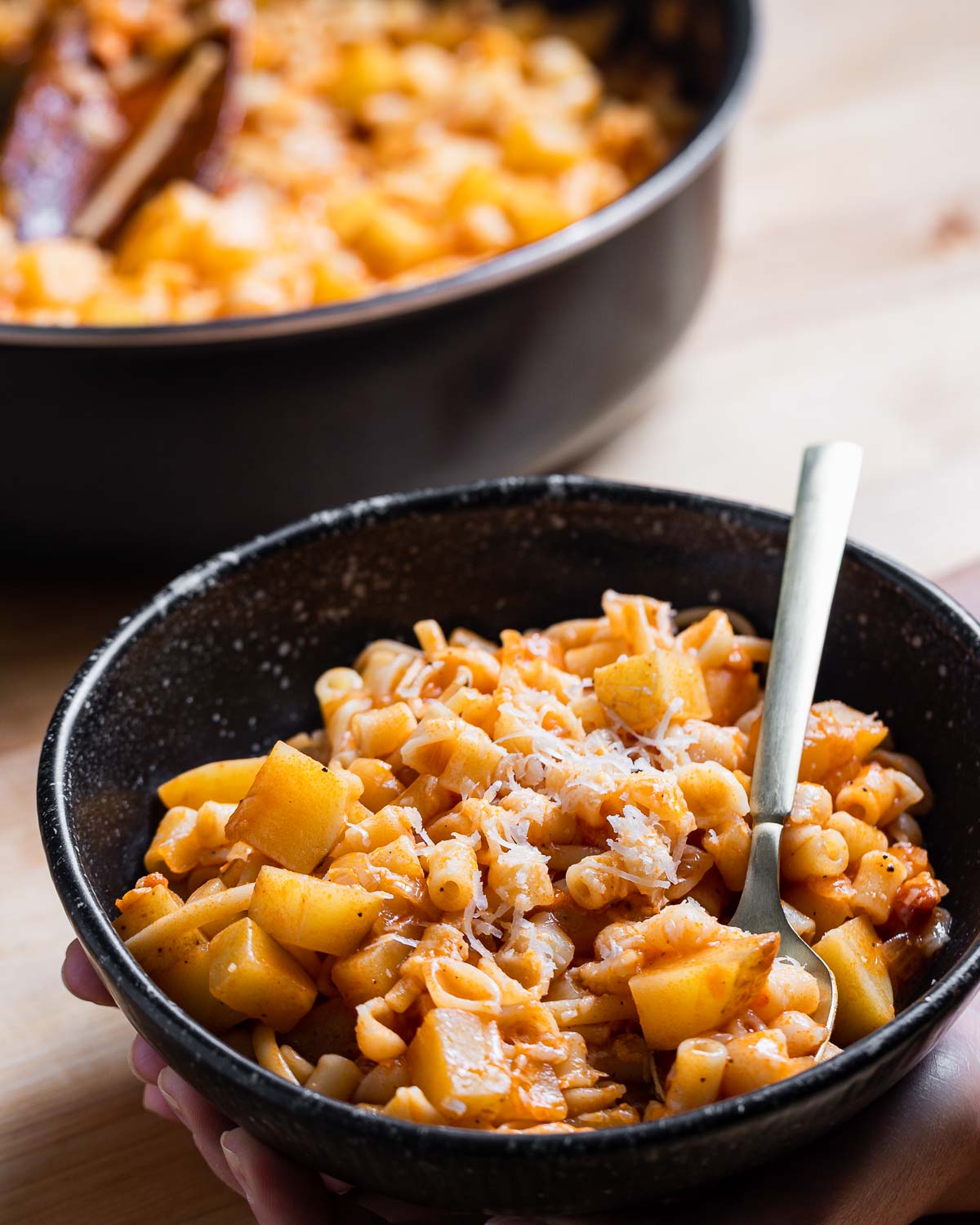 Hands holding bowl of pasta with potatoes.