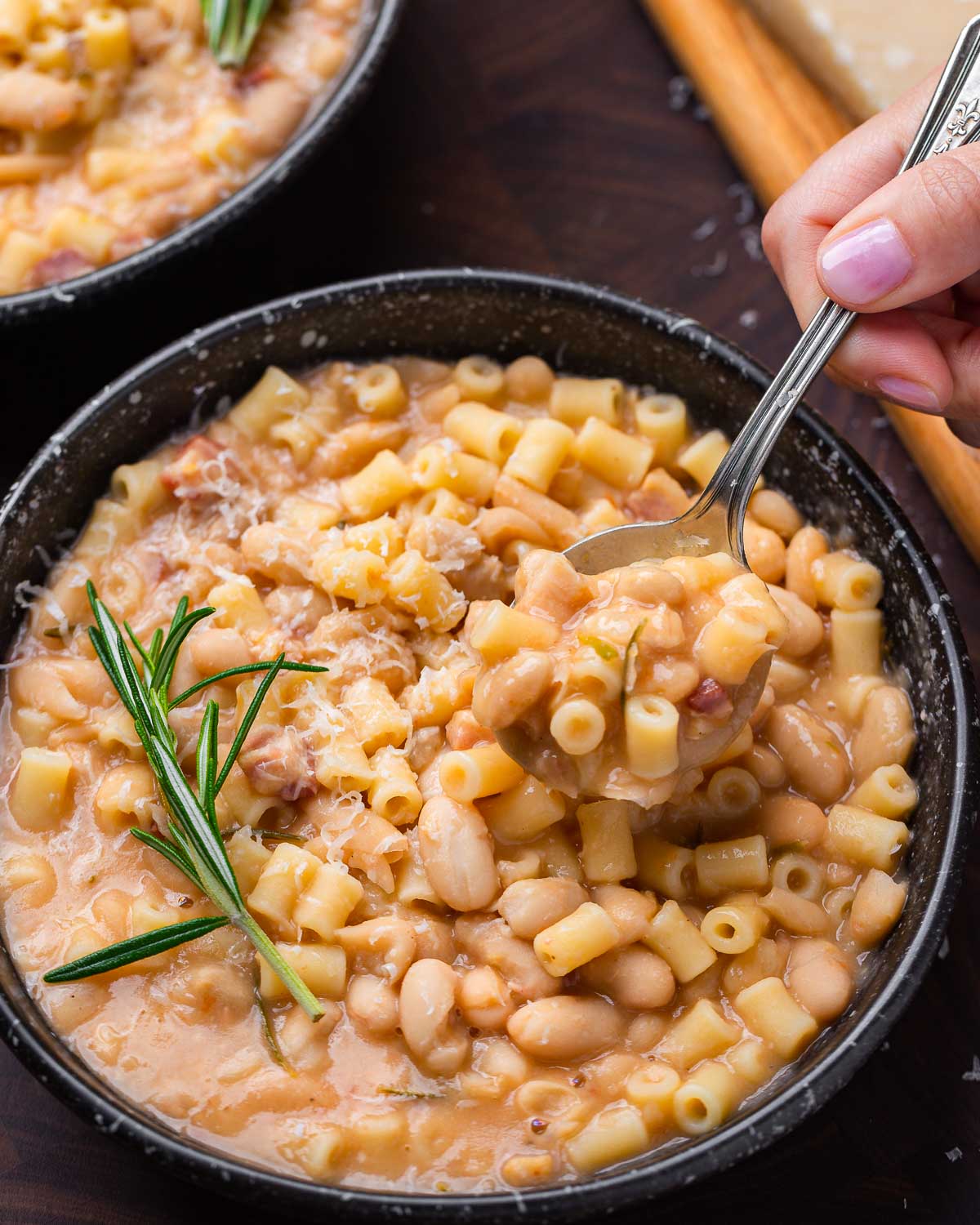 Hands holding spoonful of pasta fagioli over black bowl.