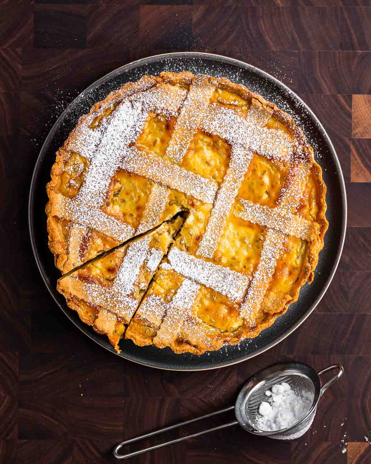 Overhead shot of pastiera Napoletana in grey plate with mesh strainer of powdered sugar.