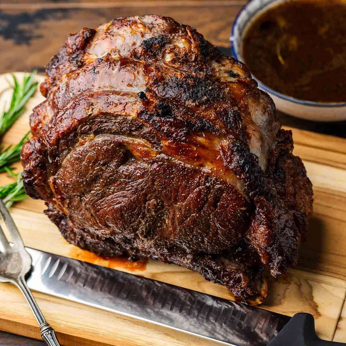 Well charred prime rib on cutting board with rosemary garnish and bowl of au jus.