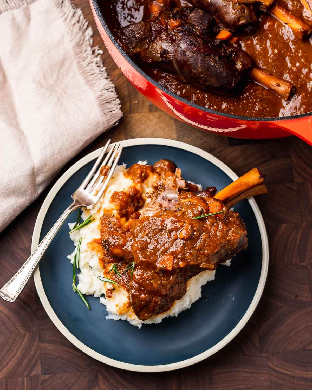 Blue plate with braised lamb shank and Dutch oven in the background.