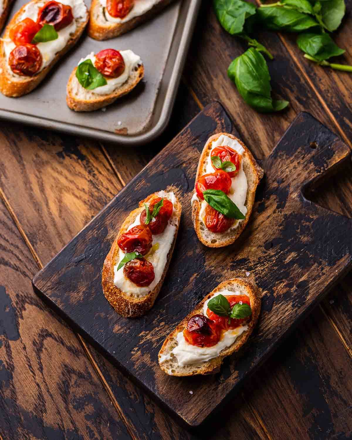 Overhead shot of ricotta roasted cherry tomato crostini on wooden serving board.