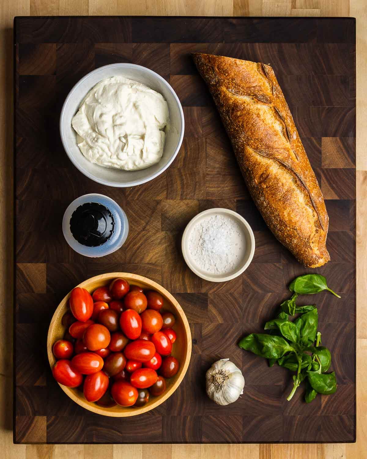 Ingredients shown: ricotta, baguette, extra virgin olive oil, flaky sea salt, cherry tomatoes, garlic, and basil.