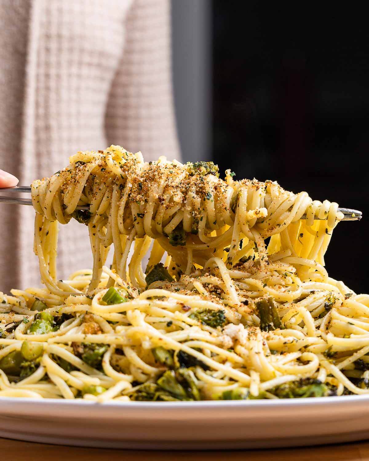 Hands holding tongs with linguine pasta topped with seasoned breadcrumbs.