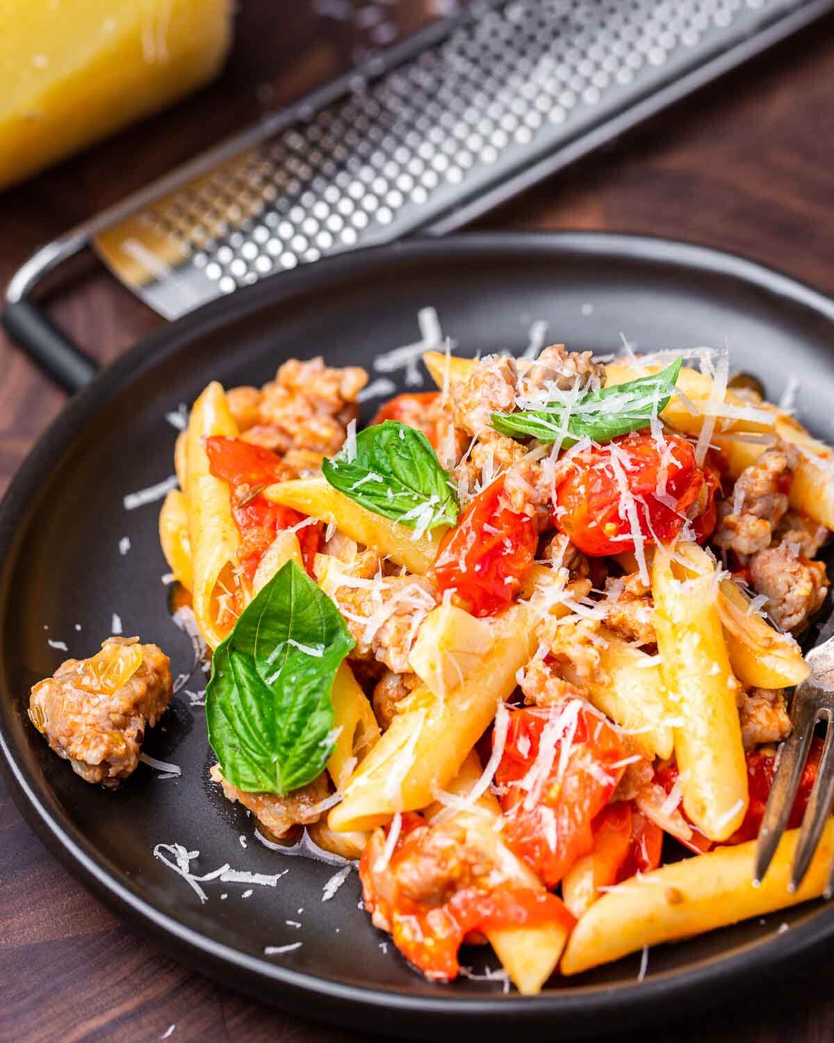 Black plate with sausage cherry tomato pasta with cheese grater in background.