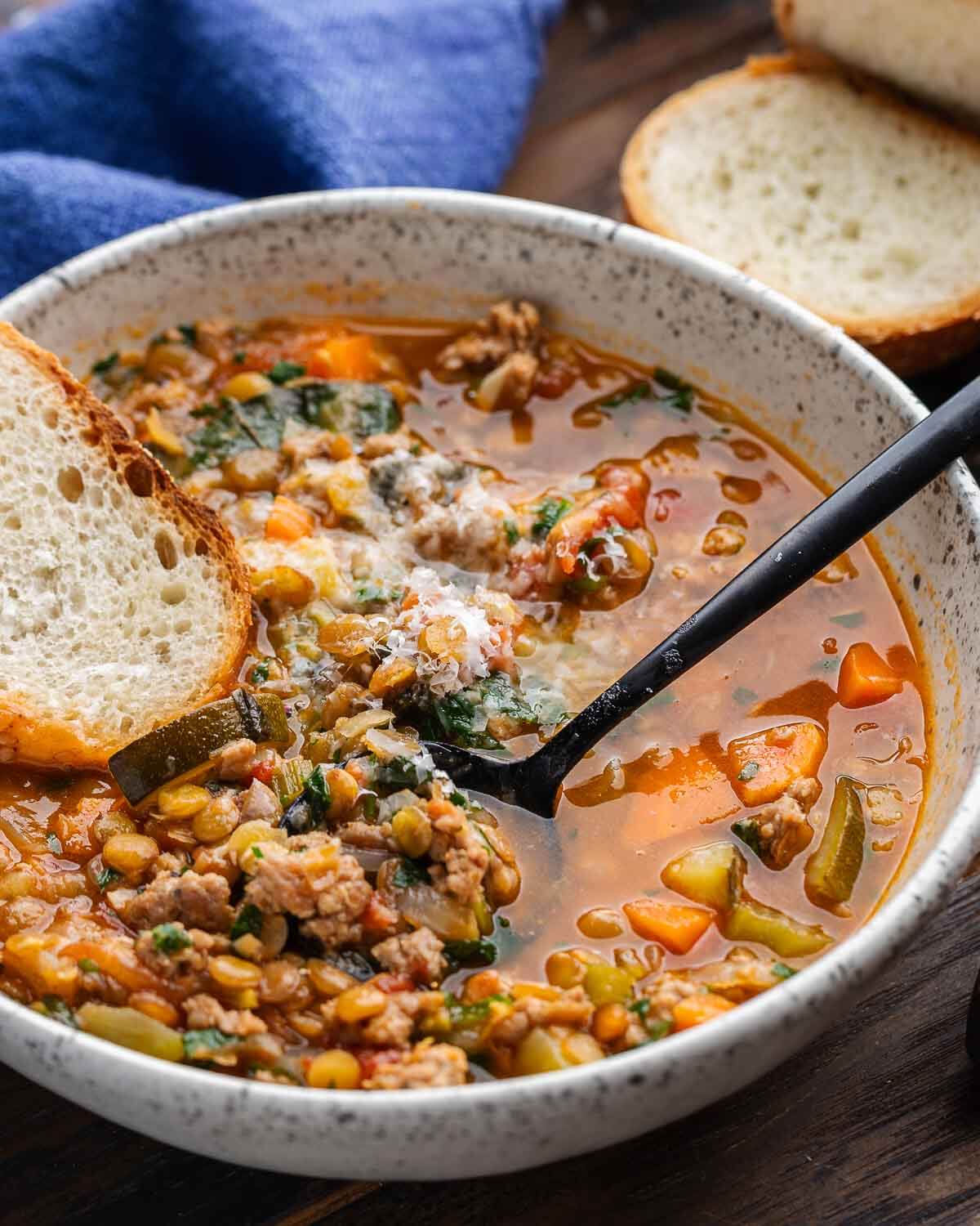 Sausage lentil soup in white bowl with slice of bread and blue napkin in the background.