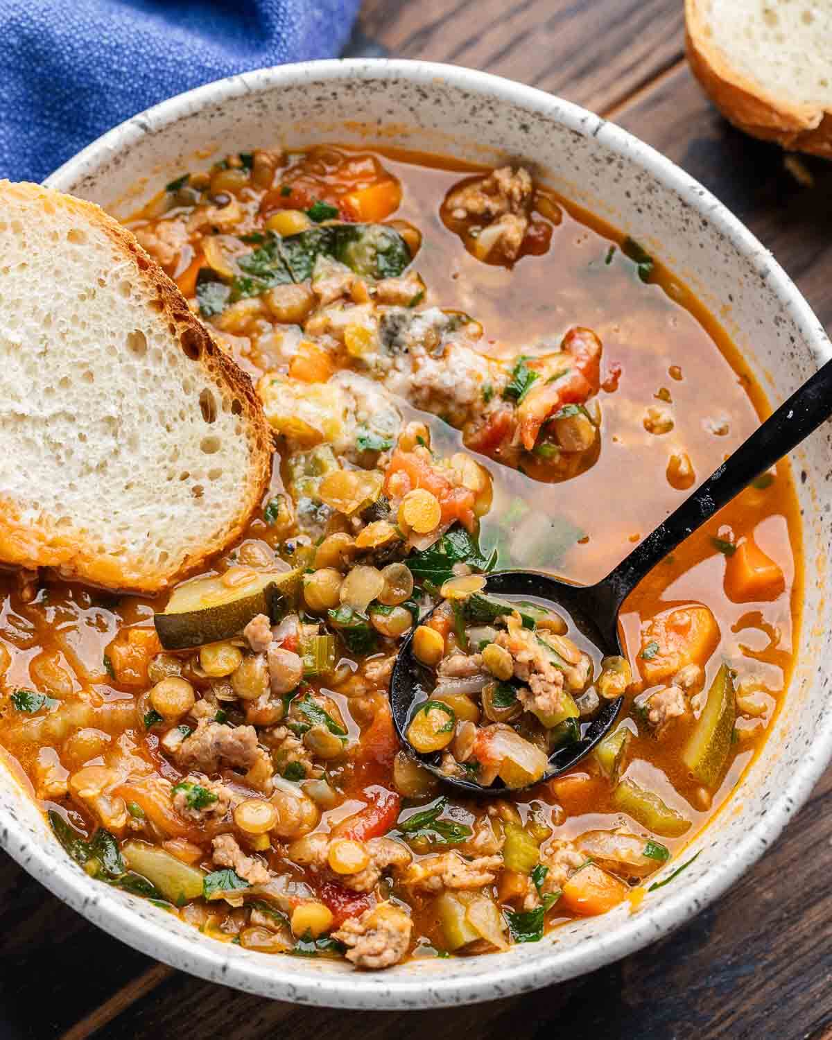 Sausage lentil soup in white bowl with slice of bread.