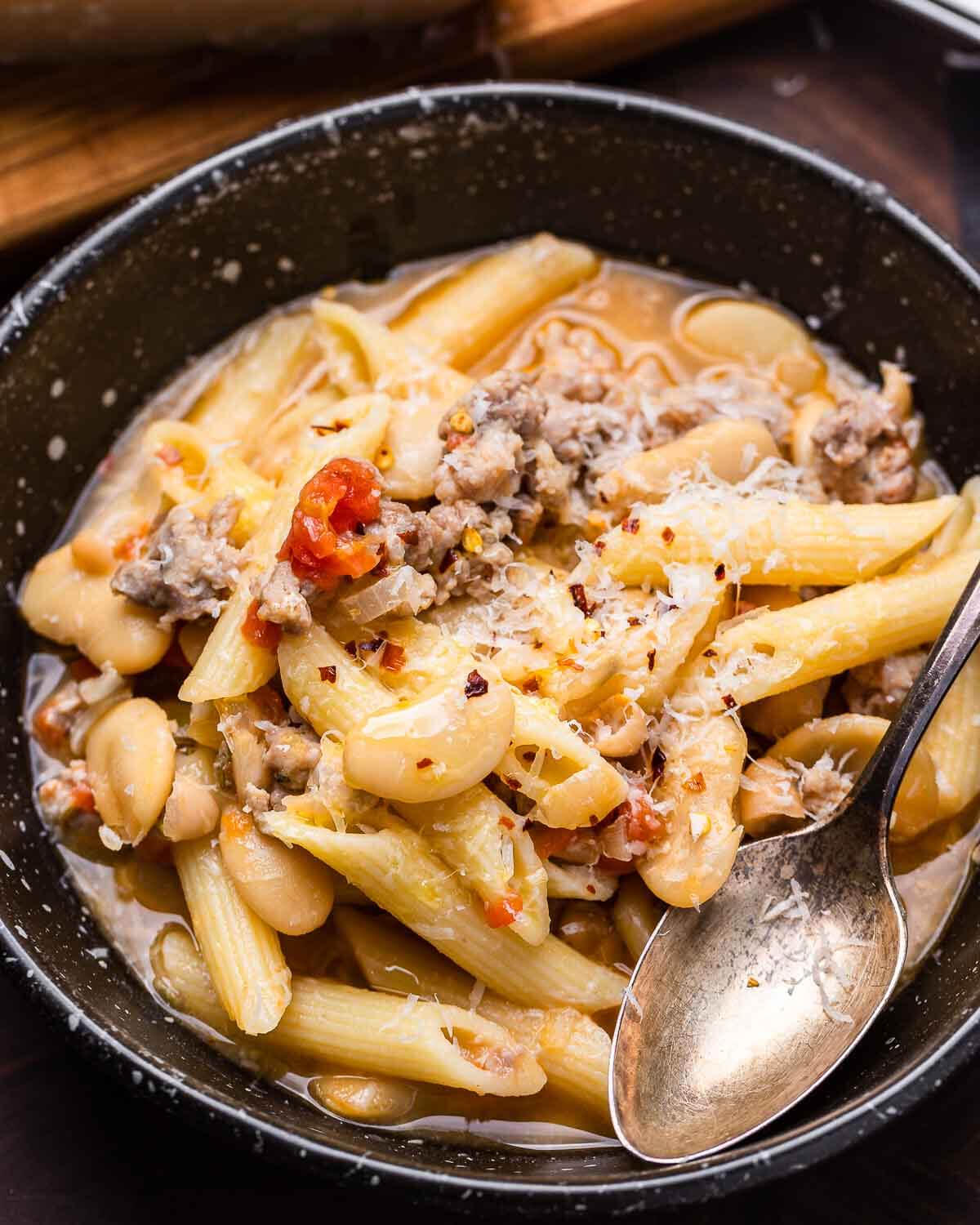 Black bowl of sausage pasta fagioli with grated cheese and spoon.