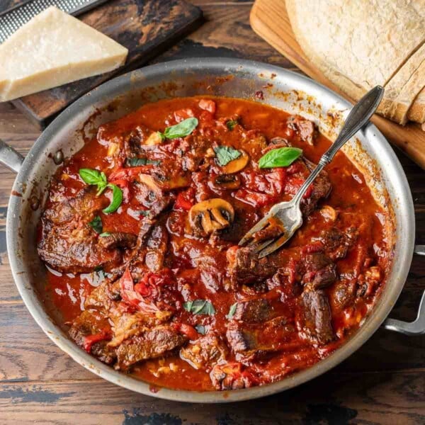 Steak pizzaiola featured image with large pan of the finished dish along with block of parmesan cheese and a loaf of bread.