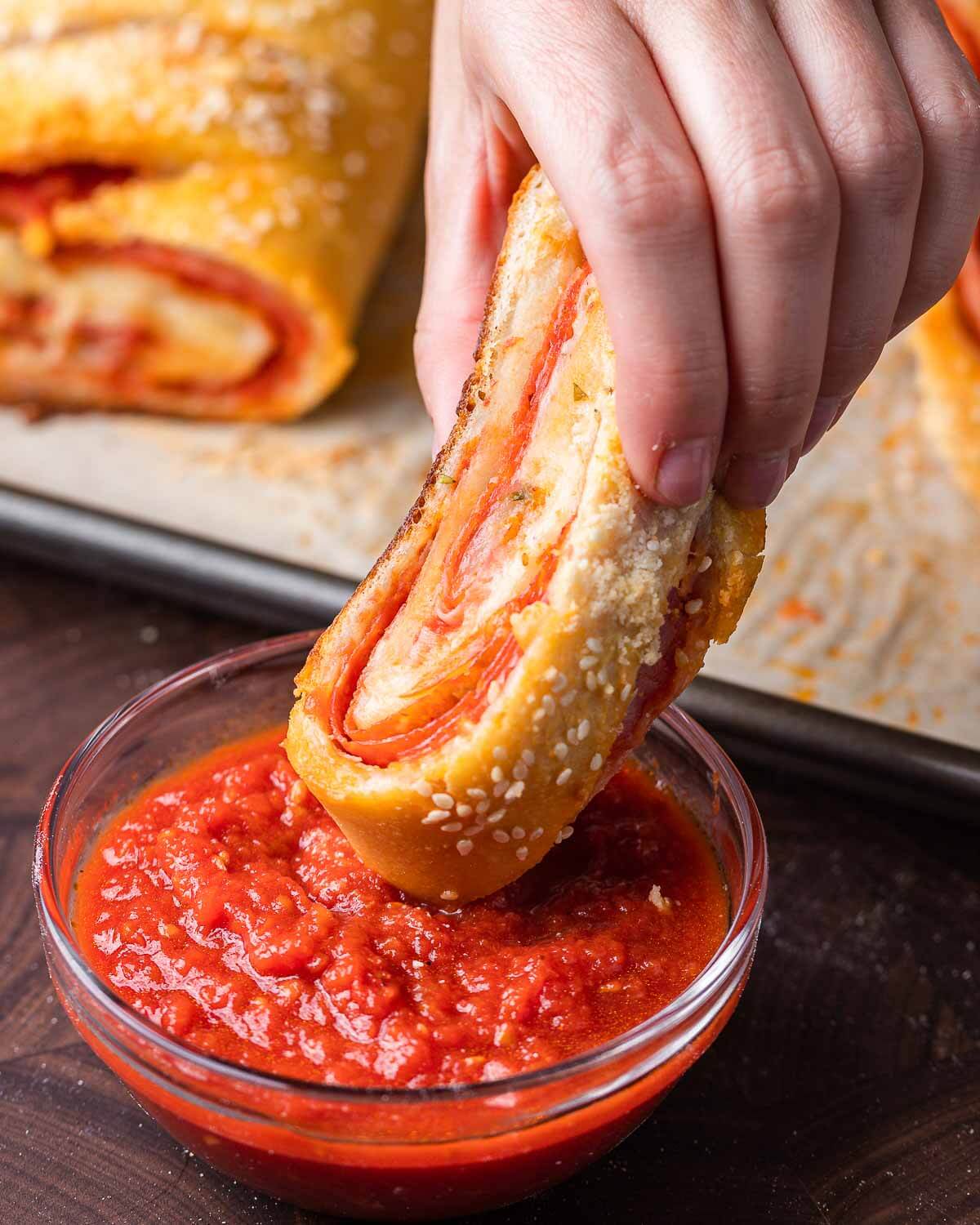 Hands dipping stromboli in marinara sauce.