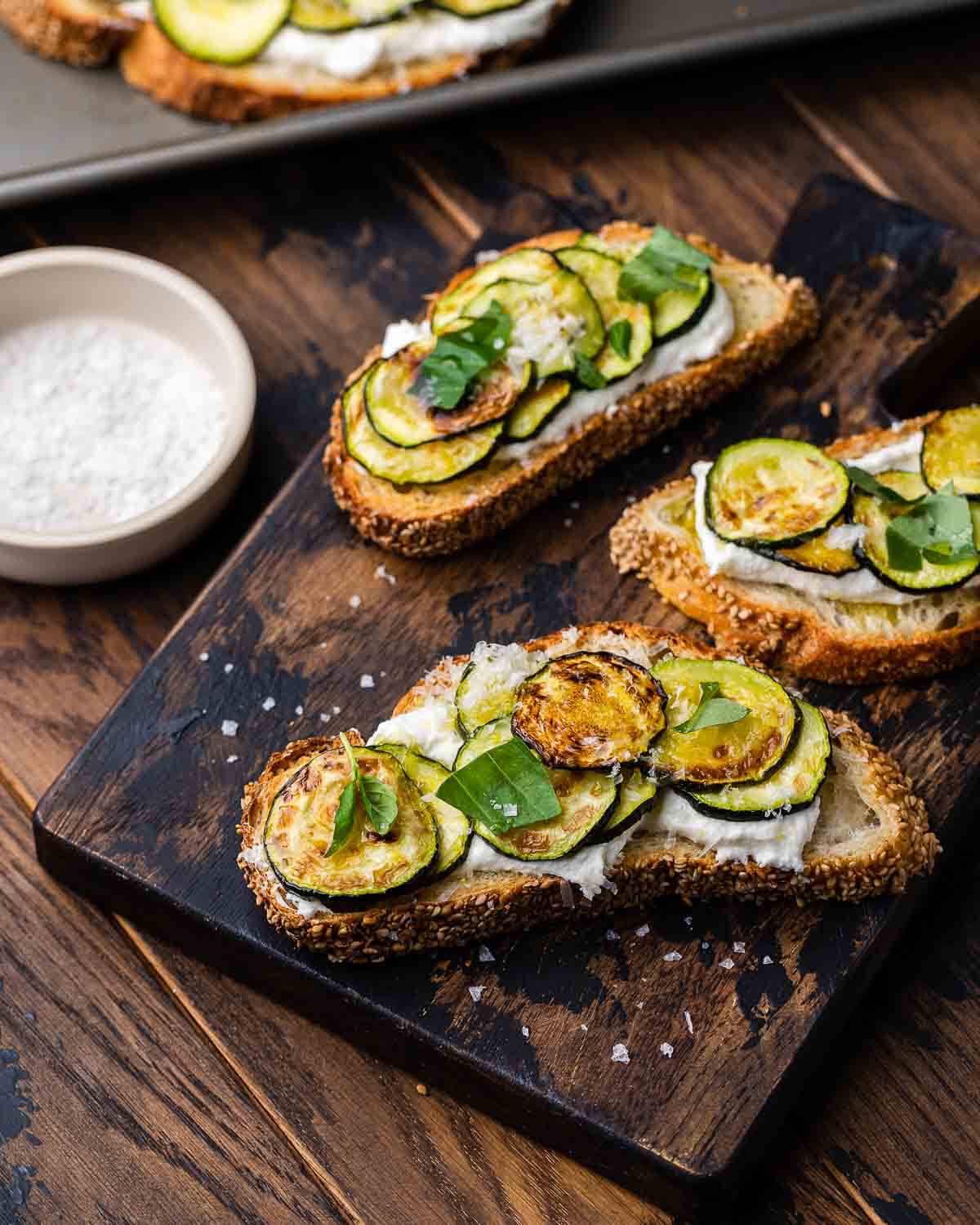 Zucchini bruschetta on wooden board with a bowl of flaky sea salt in the background.